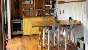 A kitchen with wooden cabinets, a yellow countertop, a stove, a microwave, and a bar stool. - Tiny house Pilolcura (Valdivia)