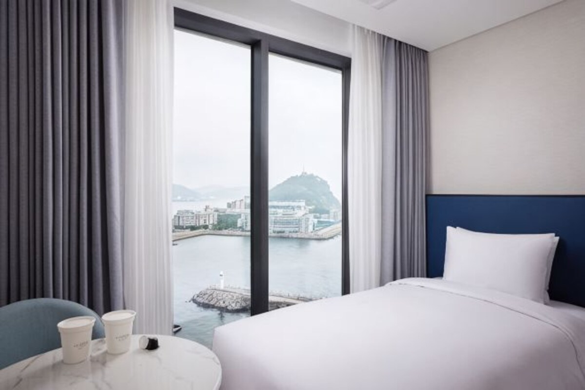Habitación de hotel con cama, una mesa redonda con tazas, vista a un cuerpo de agua y edificios, y un ventanal grande.