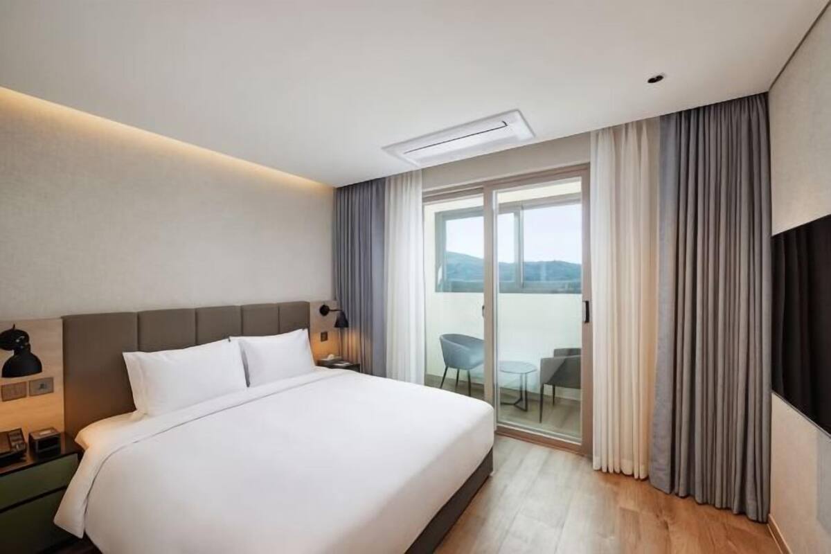 A hotel room with a large bed, a flat-screen TV, a balcony with a table and chairs, and a view of the outdoors.
