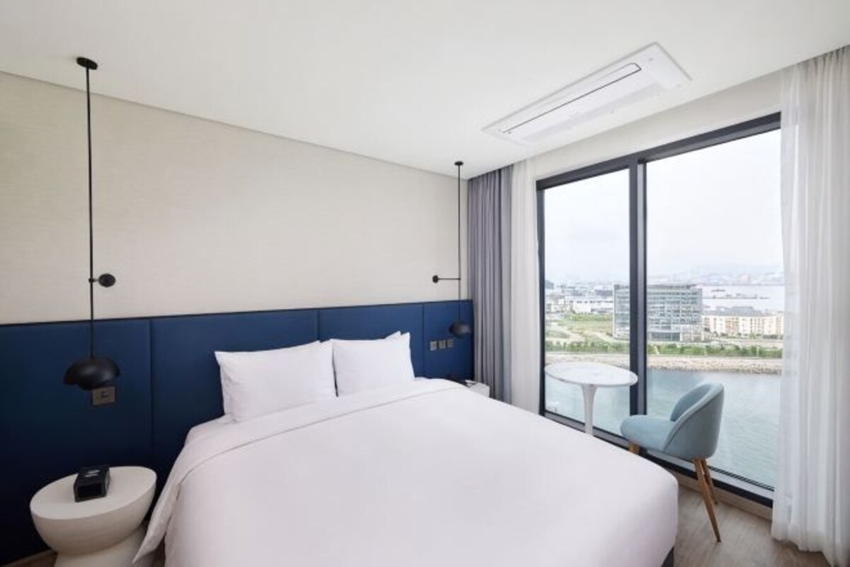 Habitación de hotel con una cama grande, una mesita de noche, una silla, una pequeña mesa y un amplio ventanal con vistas a la ciudad.