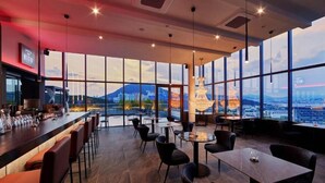 Un restaurante con zona de bar, mesas y sillas para comer, y amplios ventanales con vistas a la ciudad.