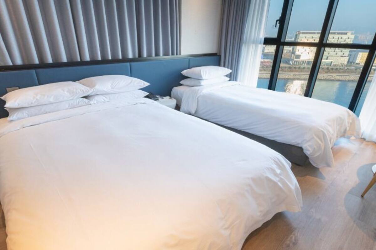 Habitación de hotel con dos camas, vistas de la ciudad a través de amplios ventanales y una pequeña mesa con una silla.