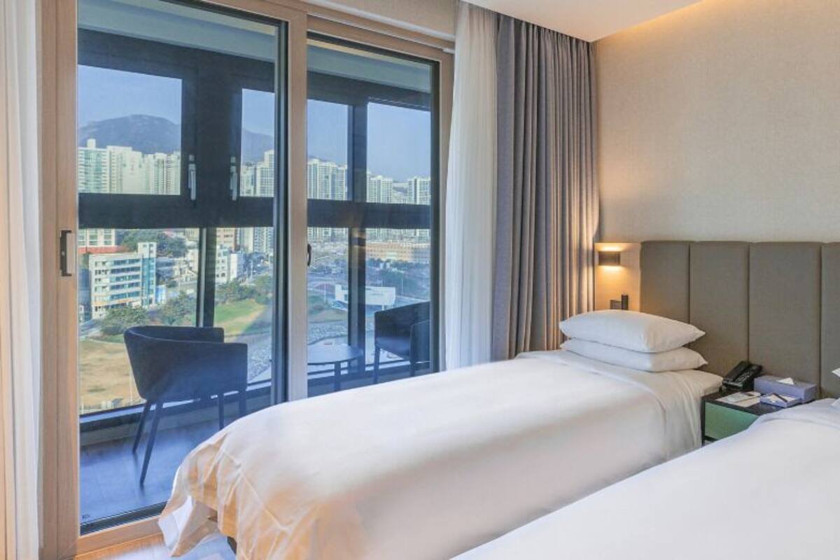 Habitación de hotel con dos camas, una mesita de noche, una silla y un ventanal grande con vistas a la ciudad.