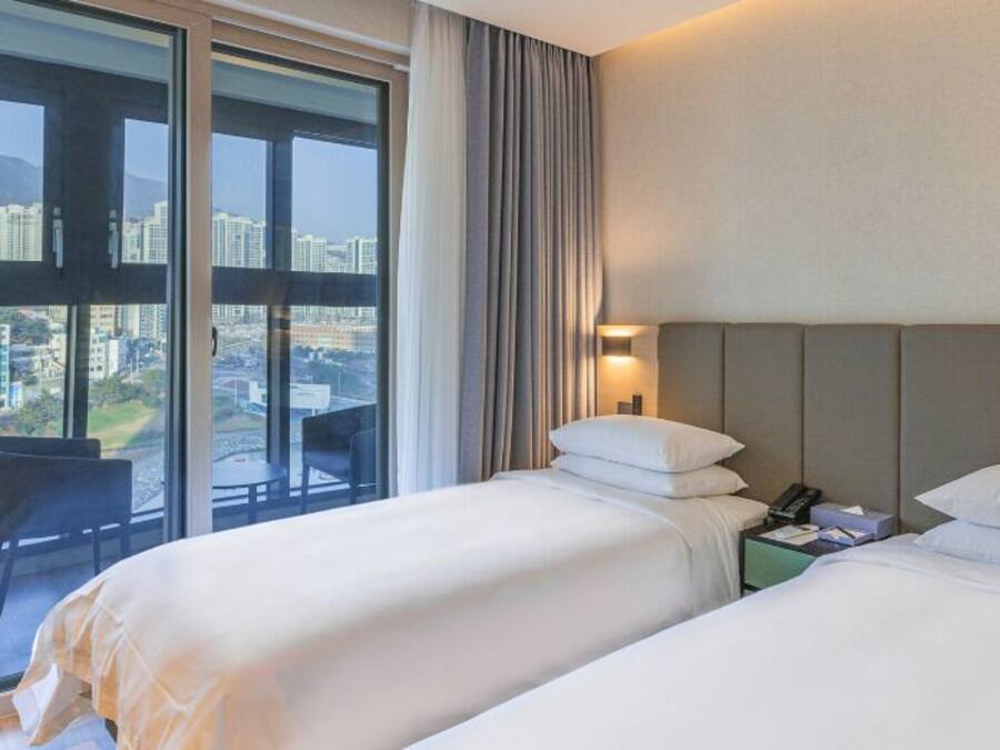 A hotel room with two beds, a nightstand, a chair, and a large window offering a city view.