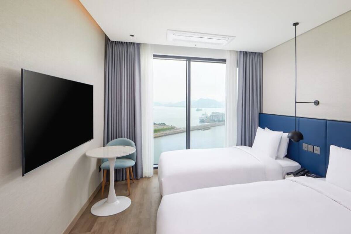 Habitación de hotel con dos camas, un televisor de pantalla plana, una pequeña mesa redonda y vista a una masa de agua a través de un ventanal grande.