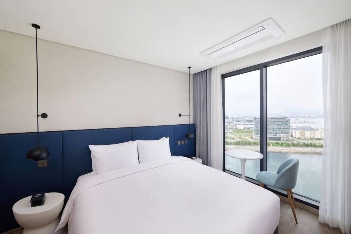 Habitación de hotel con una cama grande, una mesita de noche, una silla, una pequeña mesa y un amplio ventanal con vistas a la ciudad.