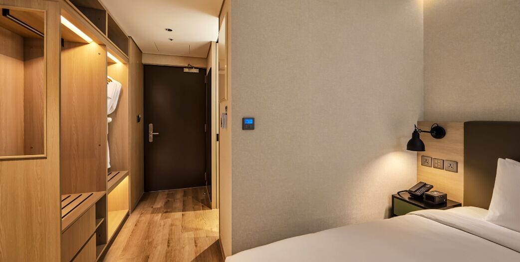 Habitación de hotel con cama, mesita de noche con lámpara, armario y puerta que da a un pasillo.