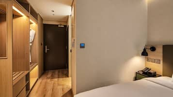 Habitación de hotel con cama, mesita de noche con lámpara, armario y puerta que da a un pasillo.