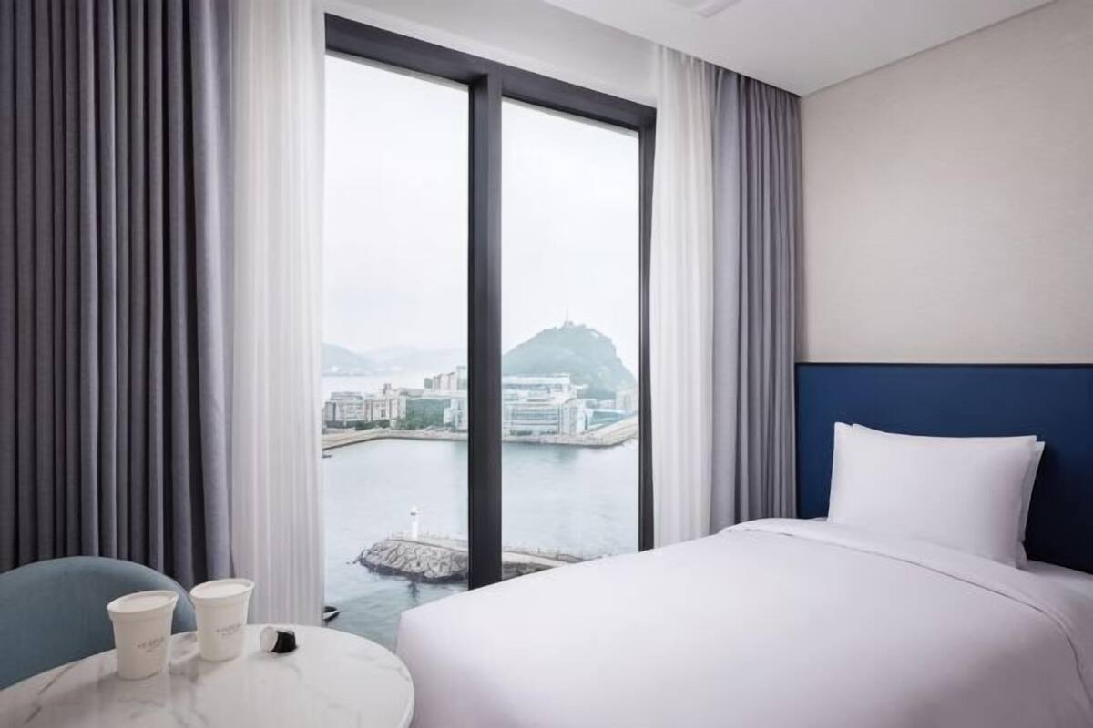 Habitación de hotel con cama, una mesa redonda con tazas, vista a un cuerpo de agua y edificios, y un ventanal grande.