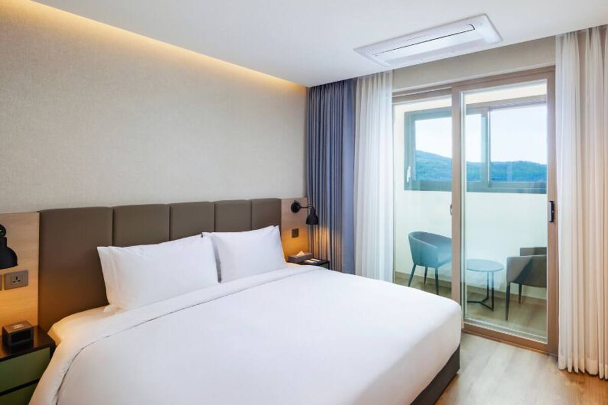 Habitación de hotel con una cama grande, una mesita de noche con lámpara, una pequeña mesa con sillas y un ventanal corredero que da a un balcón con vistas.