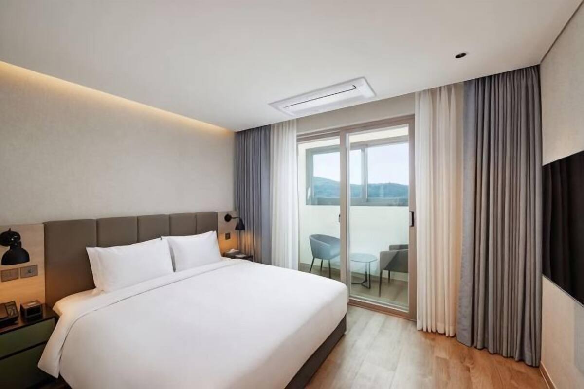 Habitación de hotel con una cama grande, televisión de pantalla plana, balcón con mesa y sillas, y vista al exterior.