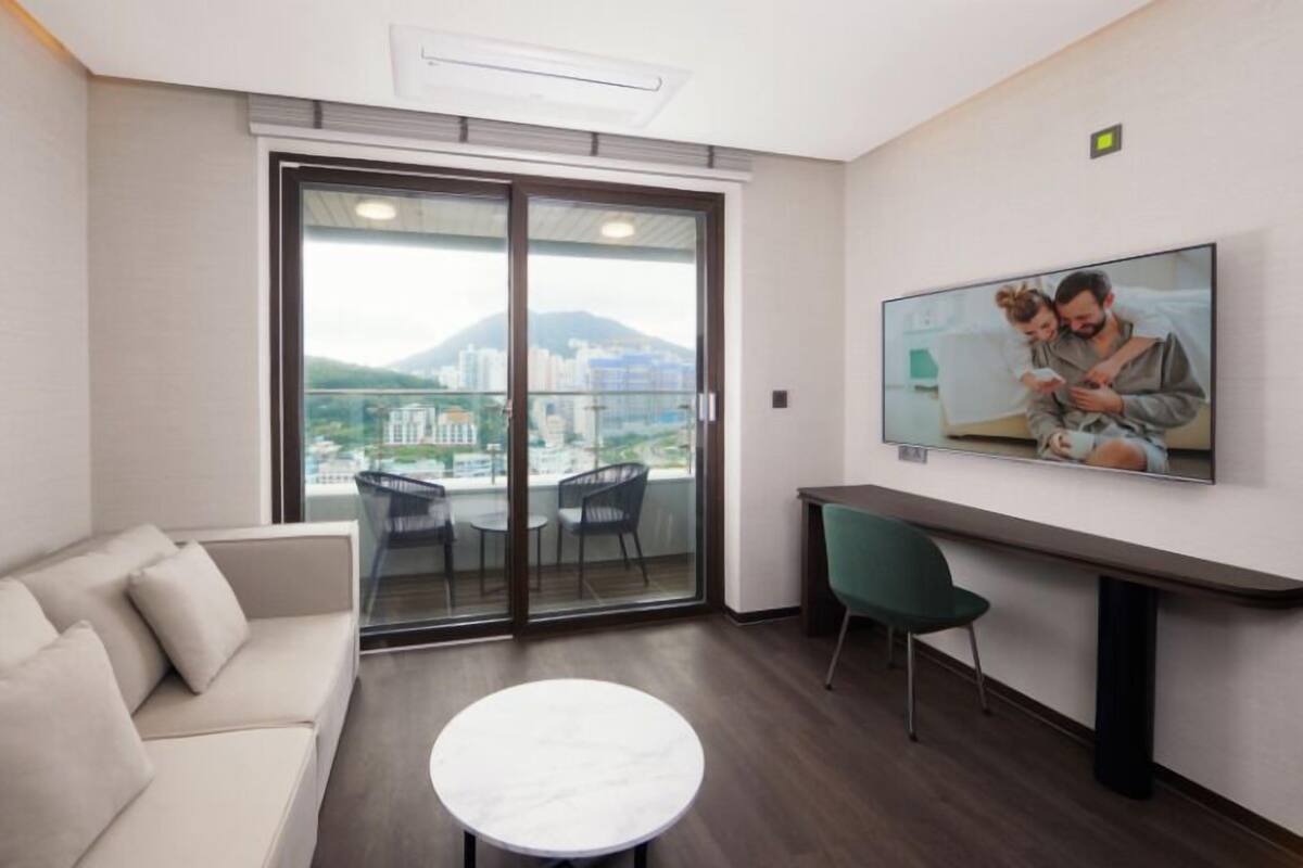 Una habitación de hotel moderna con sofá, una mesa redonda, televisor y balcón con vista a la ciudad.