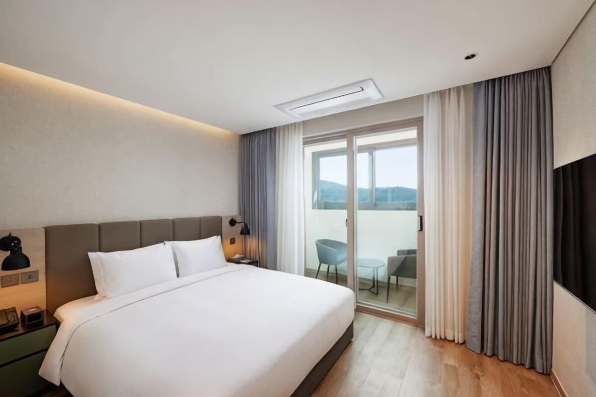 Habitación de hotel con una cama grande, televisión de pantalla plana, balcón con mesa y sillas, y vista al exterior.