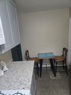 A kitchen with a granite countertop, a wooden table with a blue patterned tablecloth, two wooden chairs, and a white cabinet.