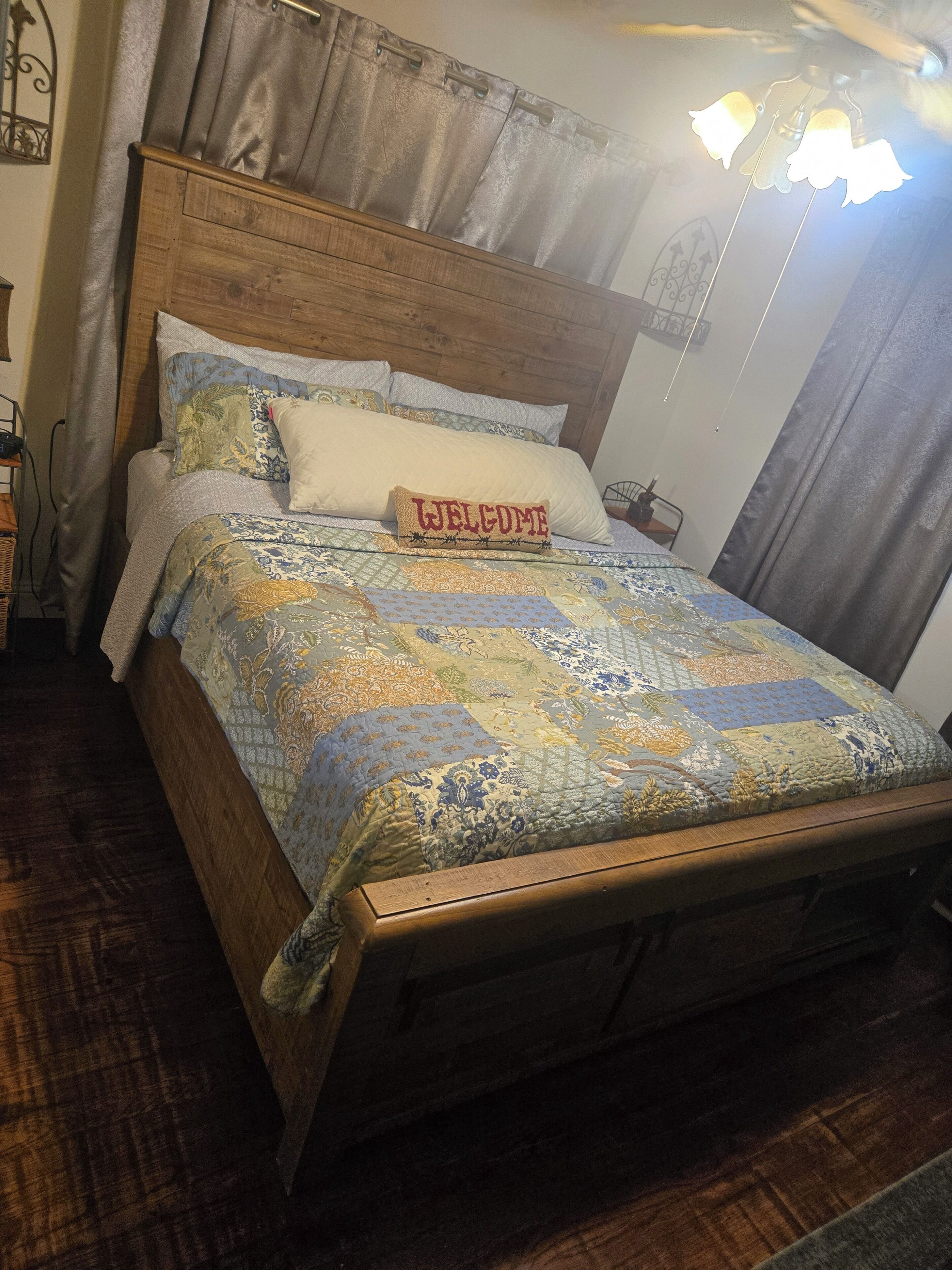 Ein hölzernes Bett mit einer gesteppten Decke, einem Kissen mit der Aufschrift 'WELCOME' und einer Nachttischlampe.