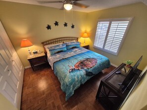 A bedroom with a bed, two bedside lamps, a ceiling fan, a window with blinds, and a small table with a plant.