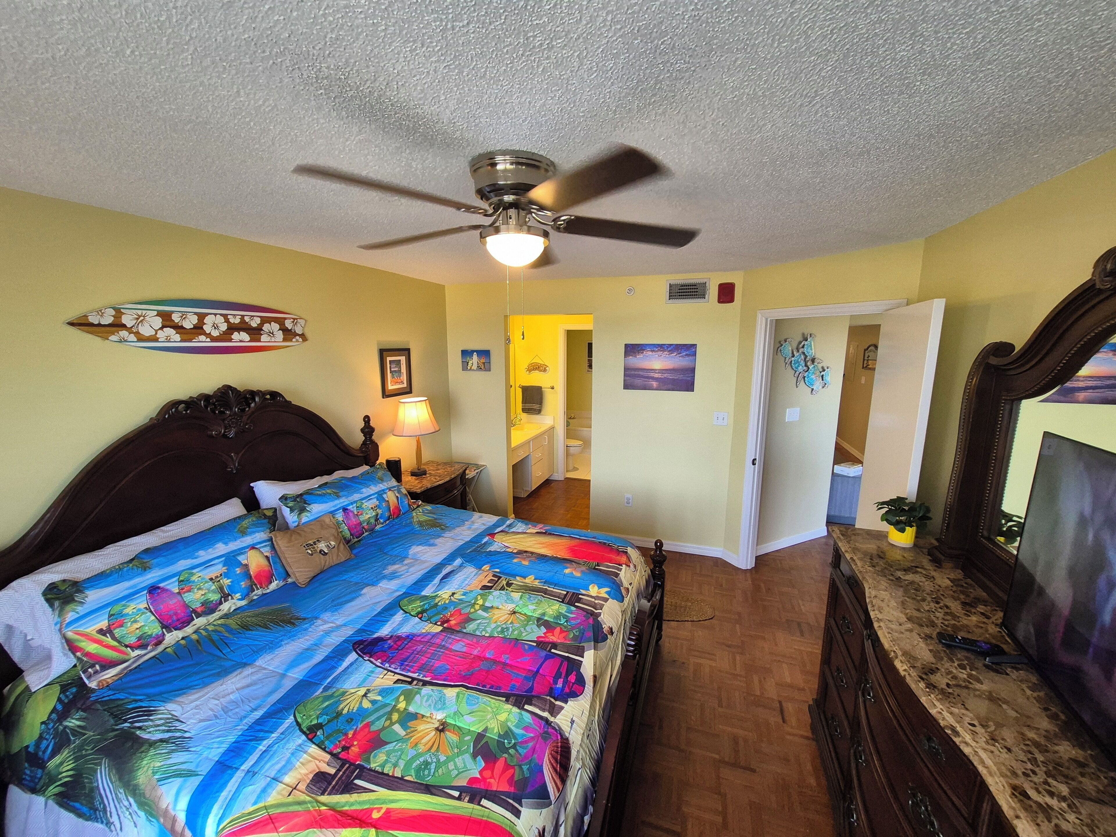 A bedroom with a bed, a ceiling fan, a nightstand, a lamp, a desk, and a TV.