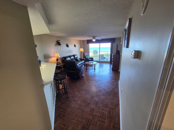 A spacious living area with a wooden herringbone floor, a black leather sofa, a dining table, and a large sliding glass door leading to a balcony with a view of the ocean.