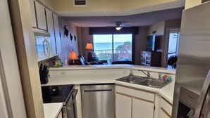 A modern kitchen with a stainless steel refrigerator, dishwasher, and sink. There is a window with a view of the ocean and a balcony with a chair.