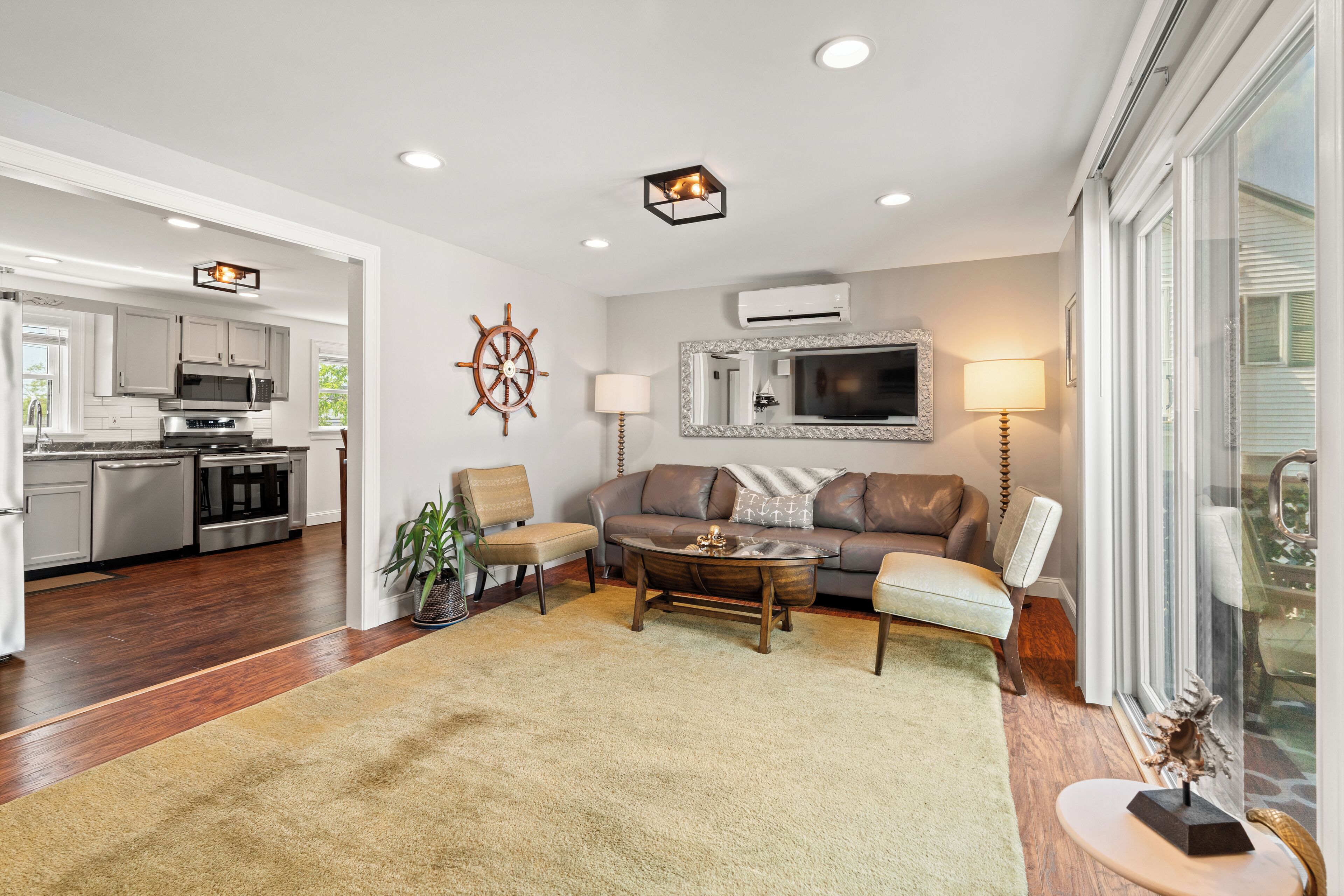 A modern living room with a sofa, coffee table, and chairs, a kitchen in the background, and a large window with a view.