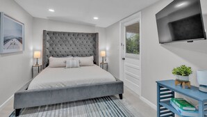 A modern bedroom with a grey upholstered bed, two bedside lamps, a flat-screen TV, a small blue side table with a plant, and a framed picture on the wall. - Walk to Lakes & Florida Southern | Pet-Friendly | 2BR, BBQ Patio, Sleeps 4 (Lakeland)