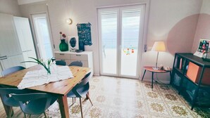 Un coin repas avec une table et des chaises en bois, une petite table d’appoint avec une lampe, un vase vert rempli de fleurs et une vue sur la mer par une porte vitrée.