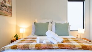 Een net opgemaakt bed met een geruit plaid, twee kussens en opgerolde handdoeken. Op het nachtkastje staan twee bedlampen en een kleine plant.
