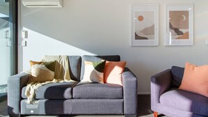 Een moderne woonkamer met twee banken, een grijze en een paarse, versierd met verschillende kussens. Twee ingelijste abstracte kunstwerken hangen aan de muur, en een raam zorgt voor natuurlijk licht.