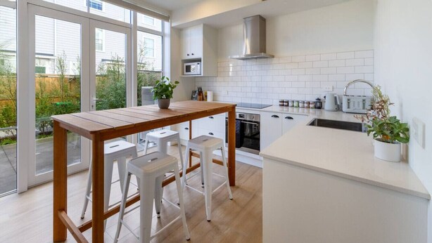 A modern kitchen with a wooden table and white stools, a microwave, oven, and a sink.