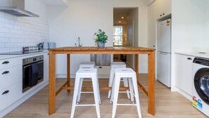 A compact kitchen with a wooden table, white stools, a built-in oven, a stainless steel range hood, and a washing machine.