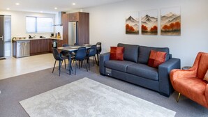 A modern living room with a grey sofa, orange armchair, and a dining area with a table and chairs. There are three paintings on the wall and a kitchen in the background. - 3bed with Garage Minutes from the Action and Sleeps 8 (Christchurch)