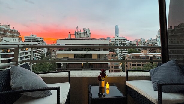 A balcony with two wicker chairs and a small table, overlooking a cityscape during sunset. - Apartment with VIP view to the mountain range, and Costanera Center. (Providencia)