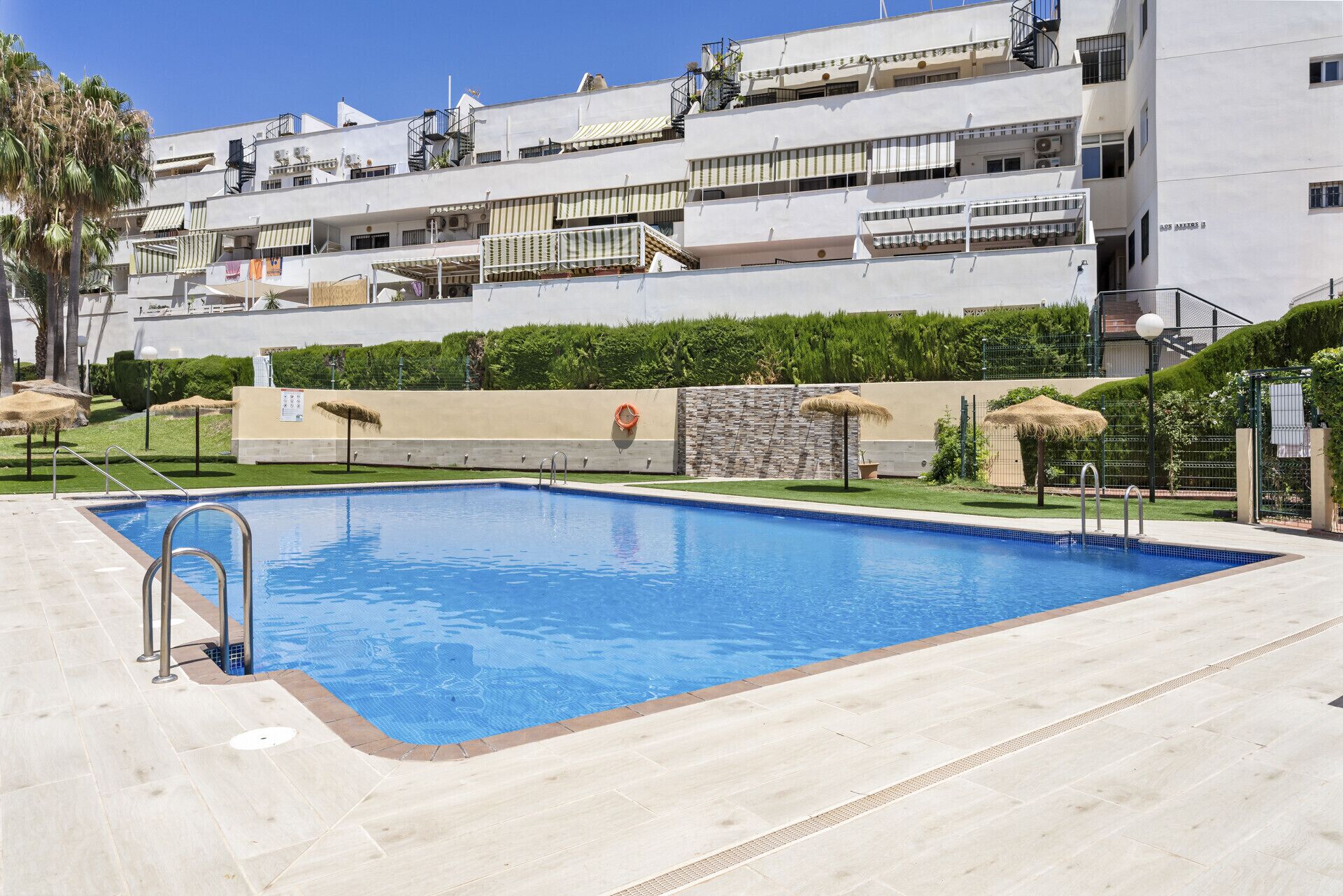 A swimming pool with a tiled deck, surrounded by a low hedge and a building with balconies.