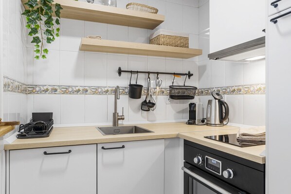 A modern kitchen with white cabinets, a stainless steel sink, a black stove, and hanging utensils. - Private Rooftop Terrace with Bar & Pool near to Torrequebrada Beach (Benalmádena)