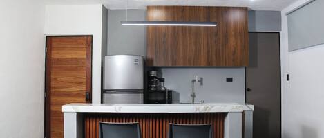 A modern kitchen with a marble countertop, two bar stools, a built-in coffee machine, and a refrigerator.