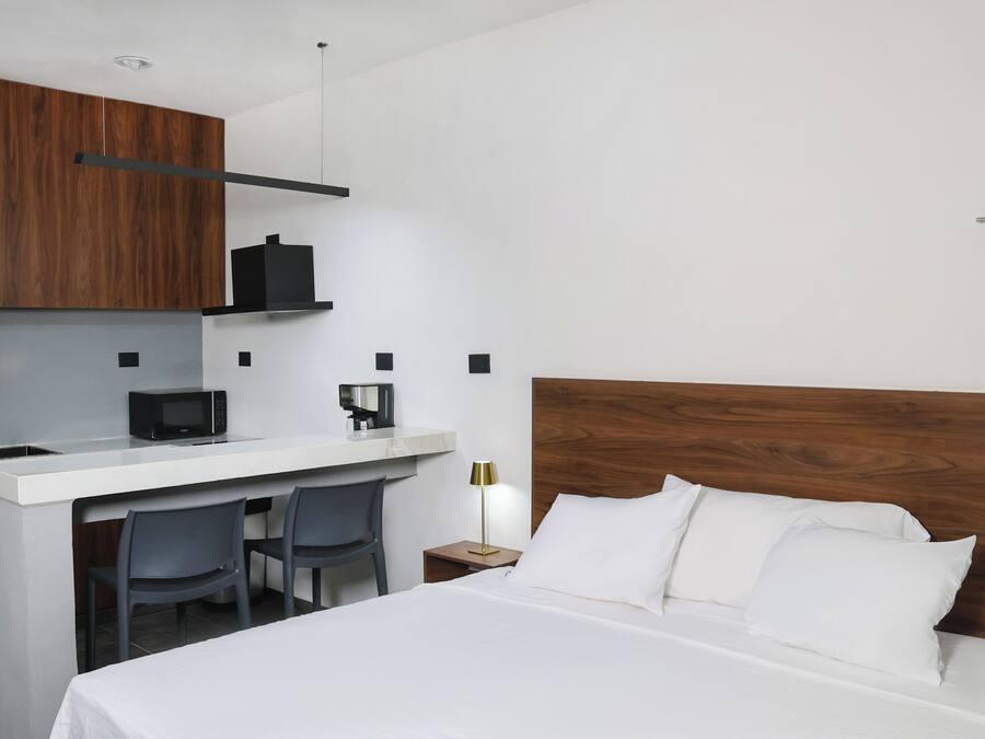 Een moderne hotelkamer met een houten hoofdeinde, een wit bed, een klein bureau met stoel, een magnetron en een nachtkastje met lamp.
