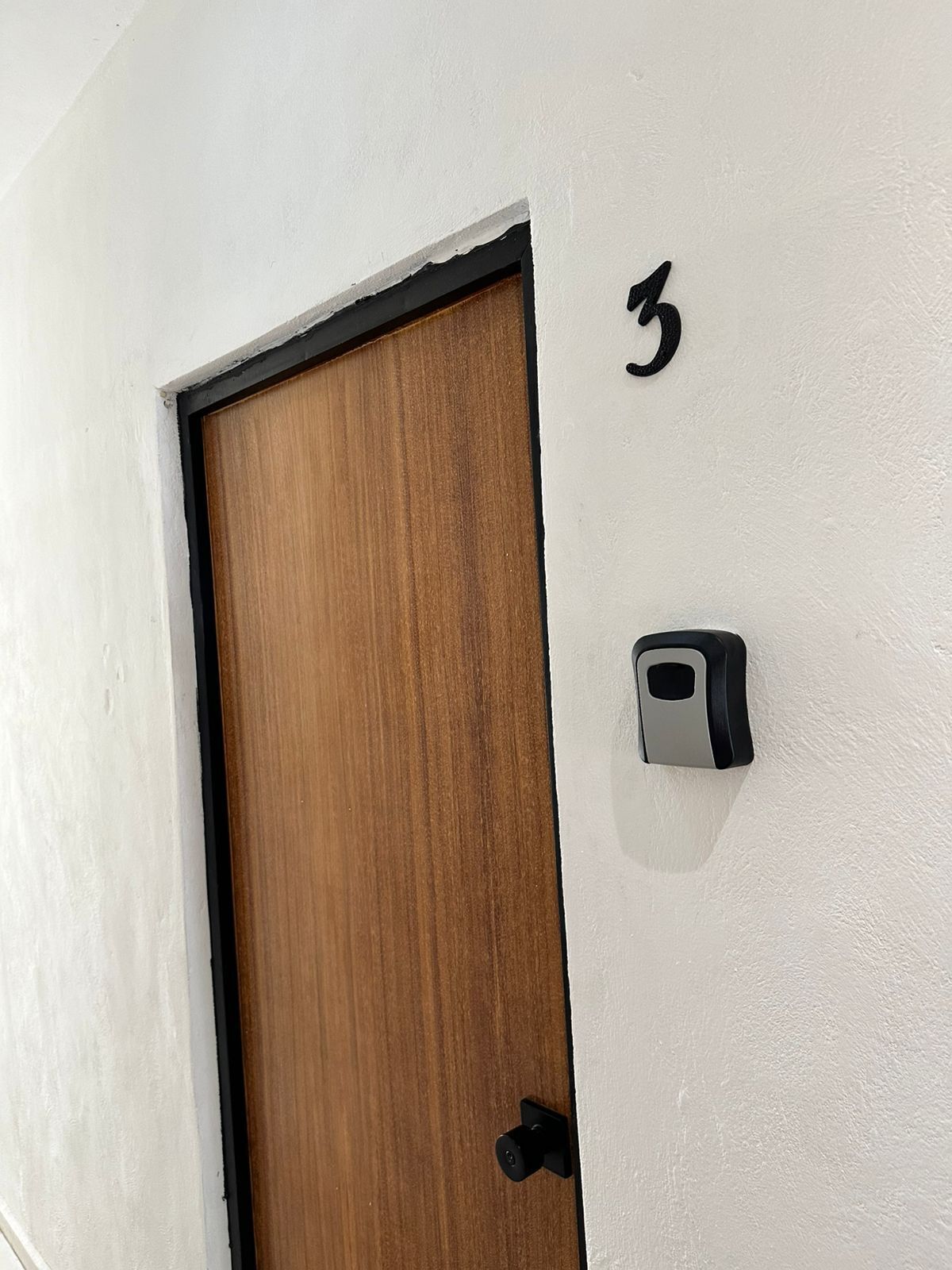 Una puerta de madera con marco negro, una pared blanca con el número 3 encima y un objeto pequeño y rectangular de color negro adherido a la pared.