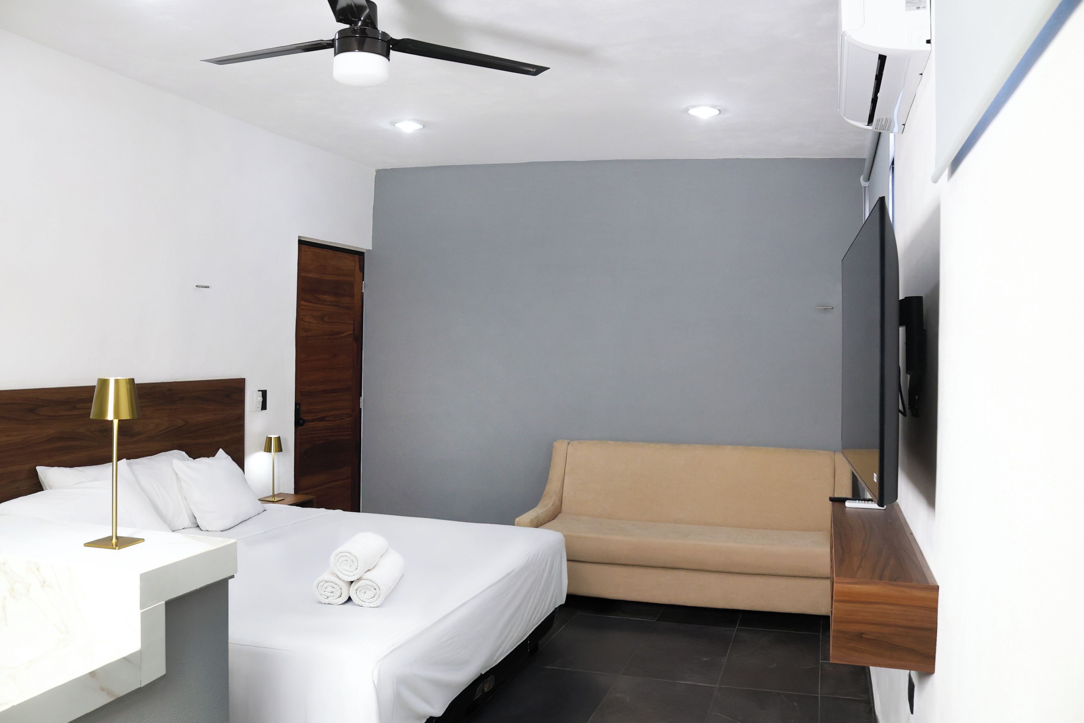 A modern hotel room with a bed, a sofa, a TV, and a ceiling fan.