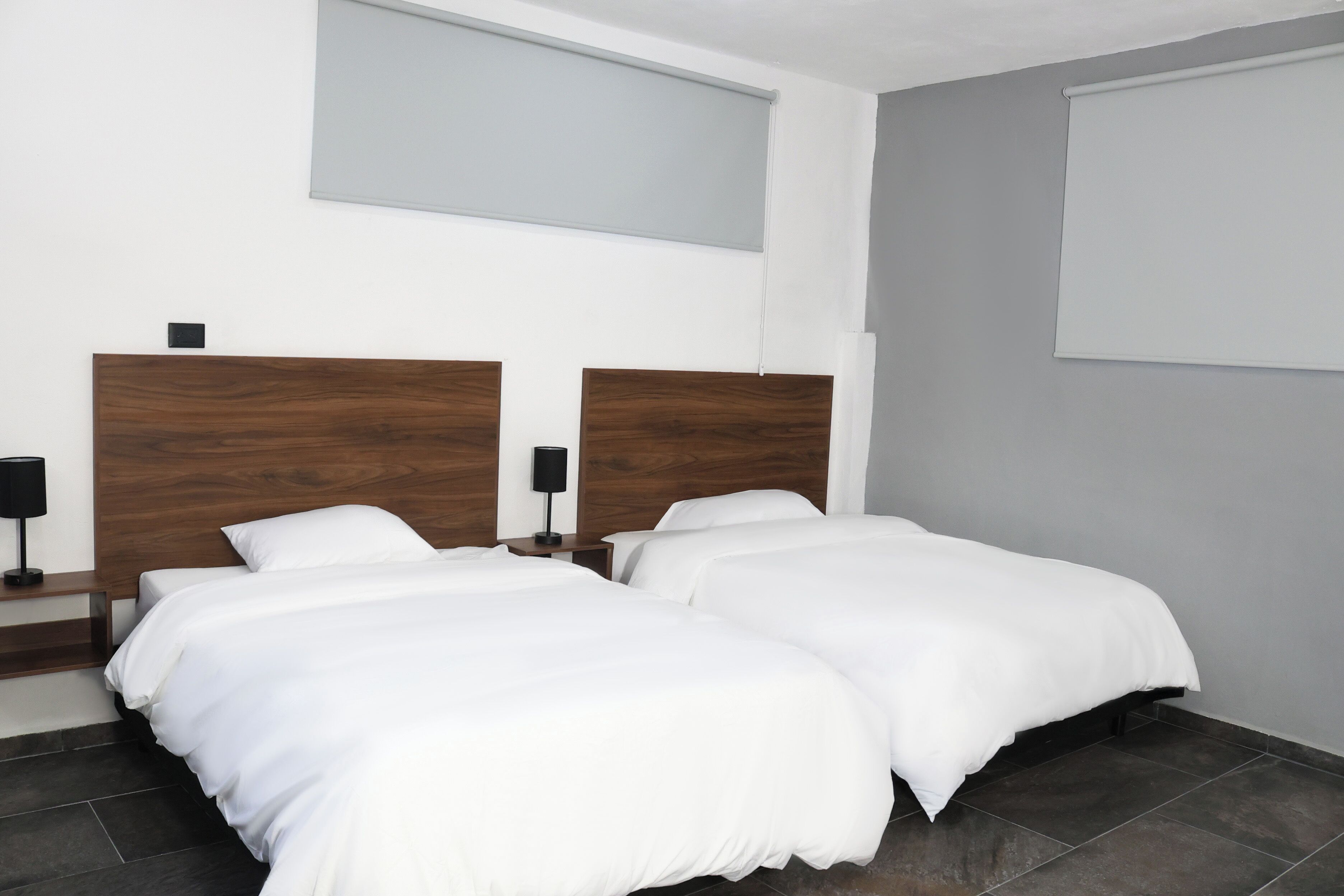 Una habitación de hotel moderna con dos camas individuales, un cabecero de madera, una mesita de noche pequeña con lámpara y un ventanal grande con persiana.