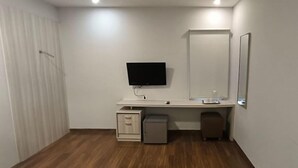 A small, clean room with a wooden floor, a white desk with a flat-screen TV, a mirror, and a small cabinet. - Hotel Bunga Bunga (Jakarta)