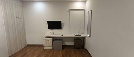 A small, clean room with a wooden floor, a white desk with a flat-screen TV, a mirror, and a small cabinet.
