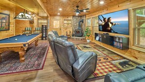 Een gezellig interieur van een chalet, met een pooltafel, leren relaxfauteuils, een open haard en een groot televisiescherm waarop twee personen te zien zijn.