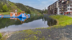 Un lac paisible, avec une structure de parc aquatique aux couleurs bleu et orange sur la gauche, entouré d’un paysage vallonné et, sur la droite, d’un immeuble à plusieurs étages doté de balcons.