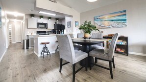 Una cocina moderna con armarios blancos, una isla central y taburetes de bar. Hay un comedor con una mesa redonda y sillas, un cuadro con una escena de playa, y una estantería con juegos de mesa.