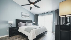 Un dormitorio con cama, mesitas de noche, ventilador de techo, ventana con cortinas y mueble para televisión.