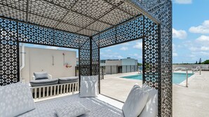 A rooftop lounge with a pool view, featuring a patterned ceiling, white cushioned seating, and a view of the pool and buildings.