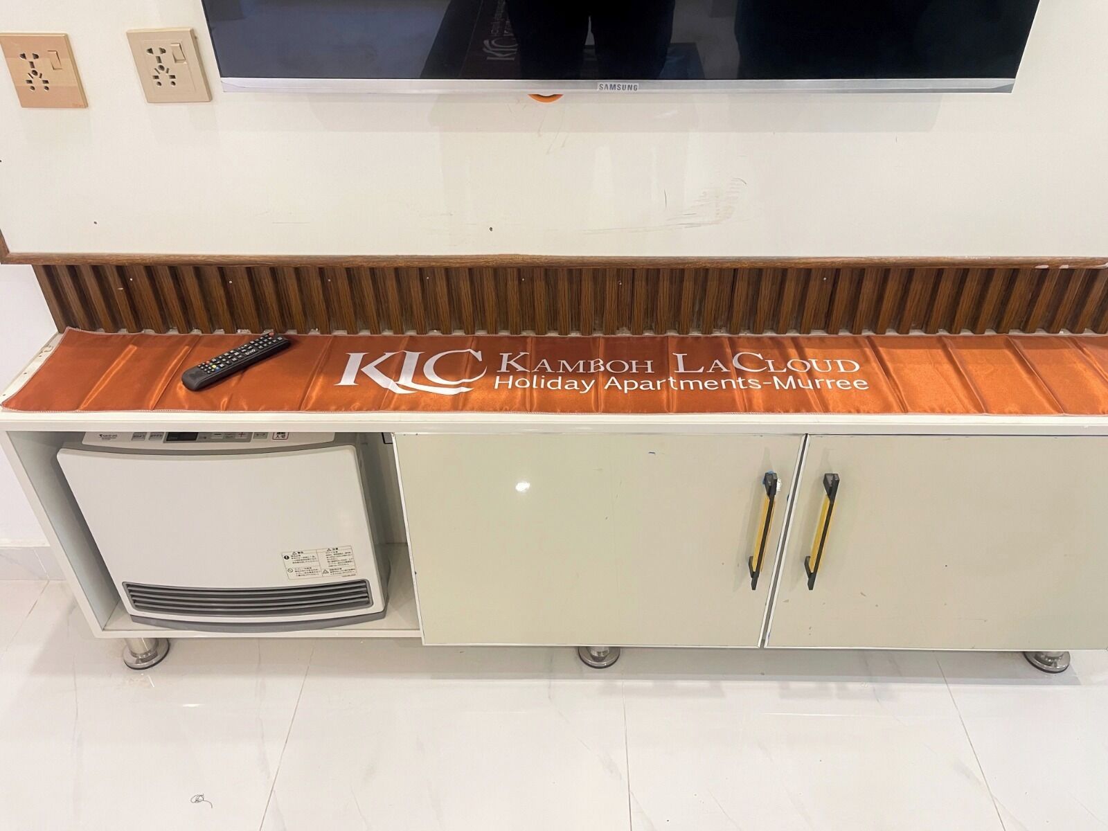 Eine weiße Minibar mit einem Fernseher darüber, einer Fernbedienung auf der obersten Ablage und einem Schild mit der Aufschrift 'KIC KAMBOH LA CLOUD Holiday Apartments-Murree'.