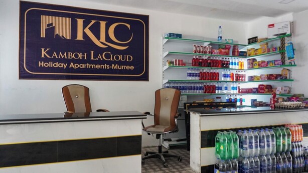Un área de recepción con un cartel que indica Apartamentos de Vacaciones Kamboh LaCloud-Murree, que incluye un mostrador de recepción, dos sillas y estantes repletos de bebidas y aperitivos.