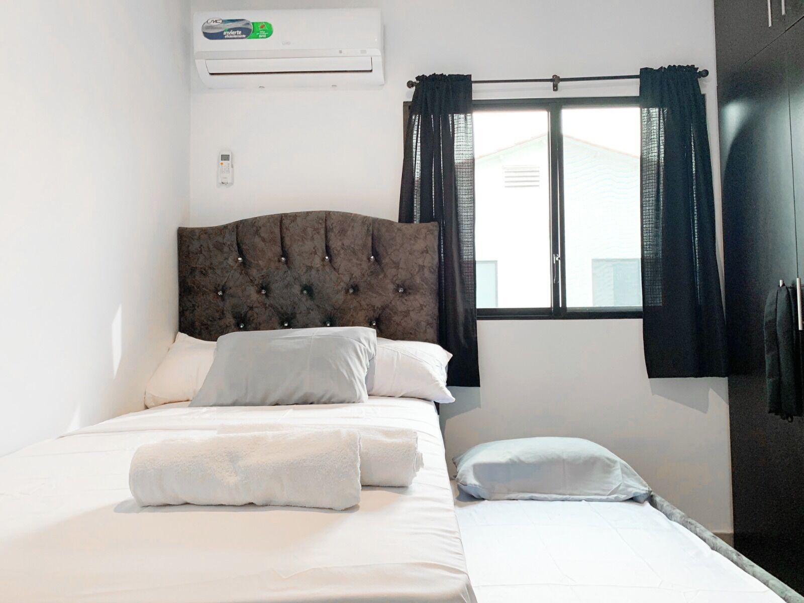 A bedroom with a bed, a window with curtains, an air conditioning unit, and a wardrobe.