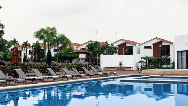 Una piscina con sillas de descanso y sombrillas, rodeada de edificios y follaje.